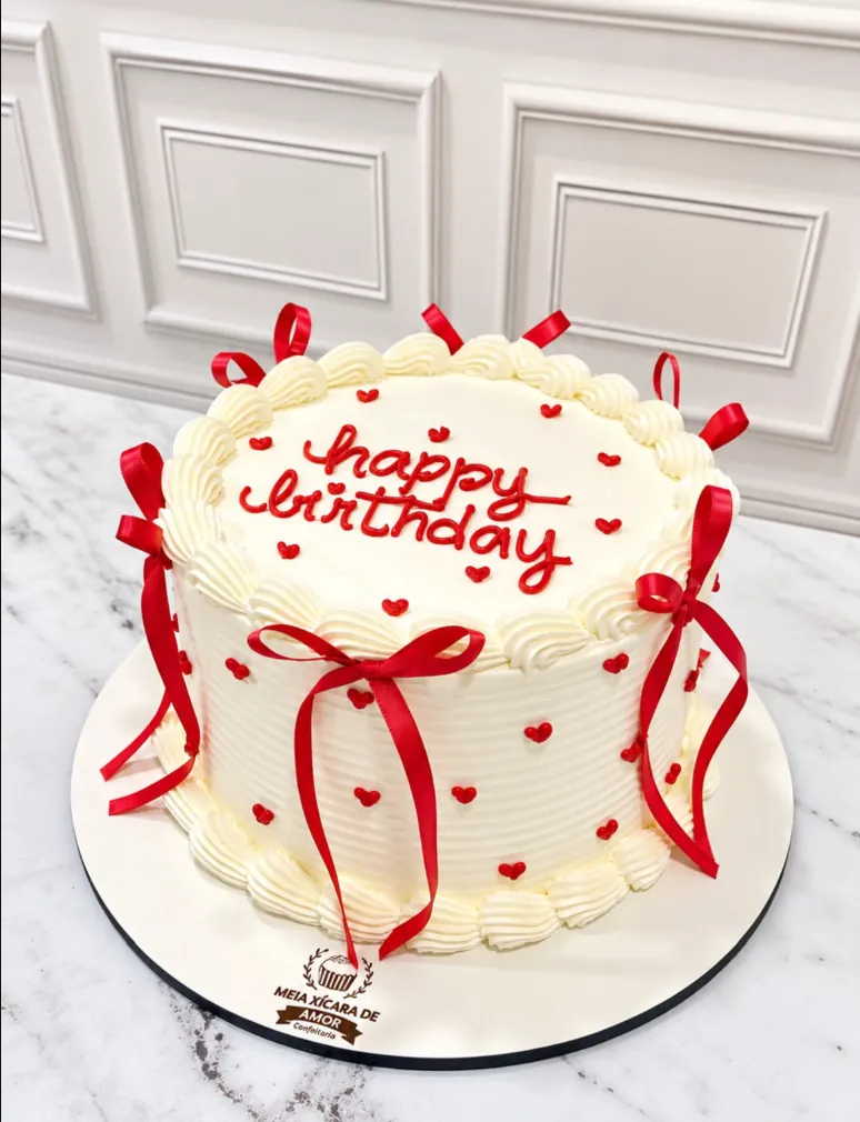 Bolo branco com 'happy birthday' em vermelho, corações e laços de fita vermelha